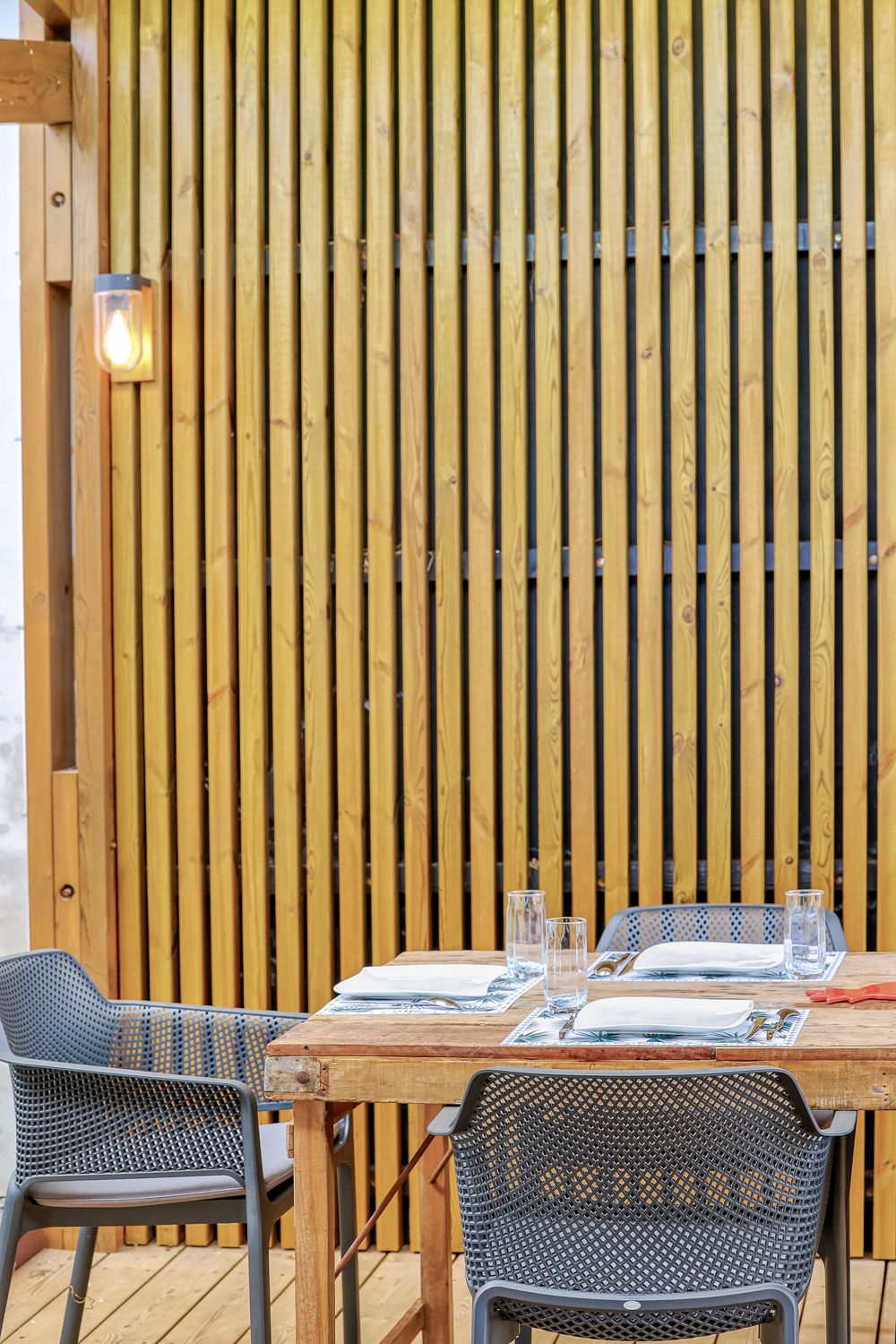 Rustic wooden dining table with gray chairs against vertical wood slat wall with wall sconce lighting.