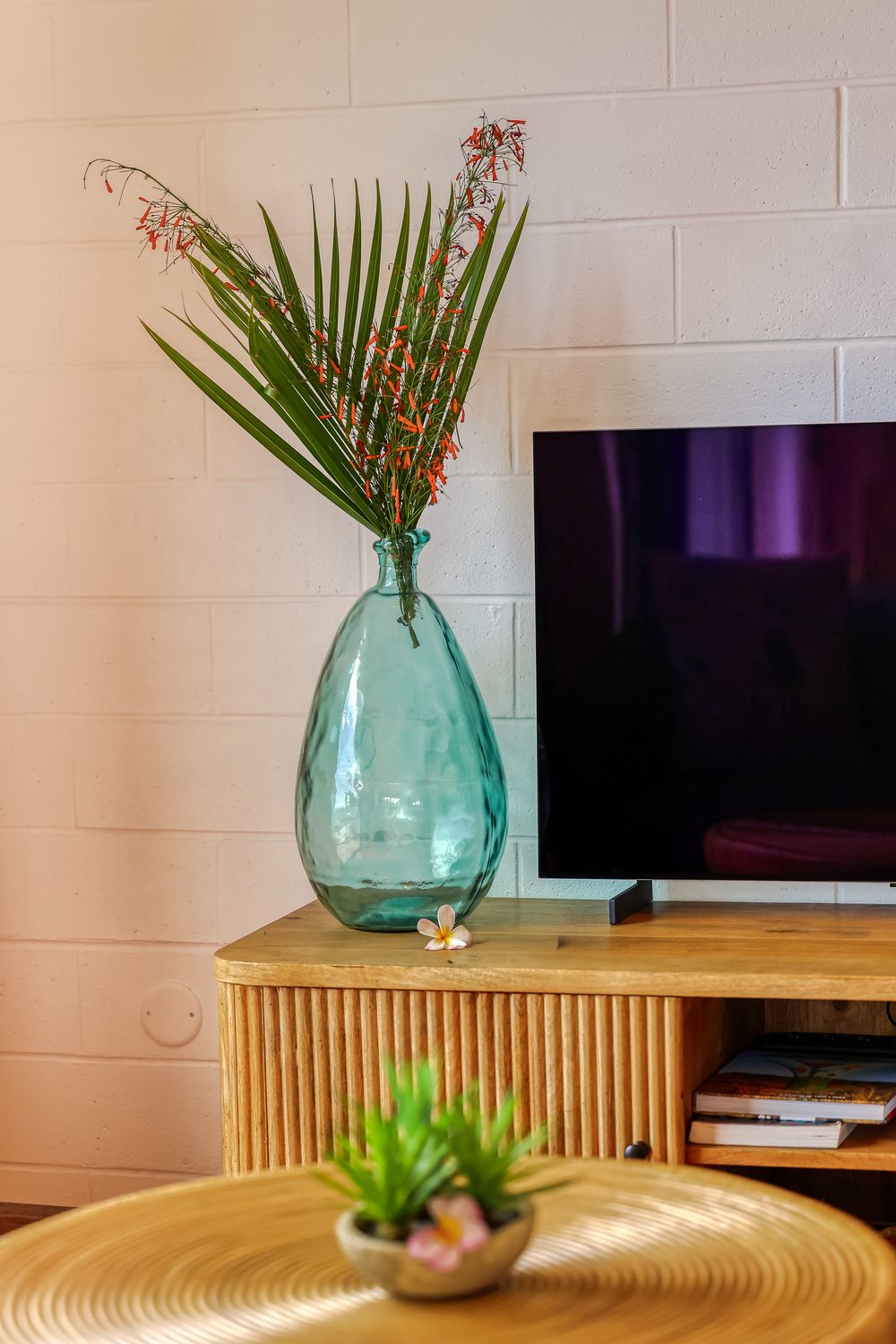 A teal glass vase with tropical palm leaves and red flowers on a wooden TV console beside a flat-screen television.