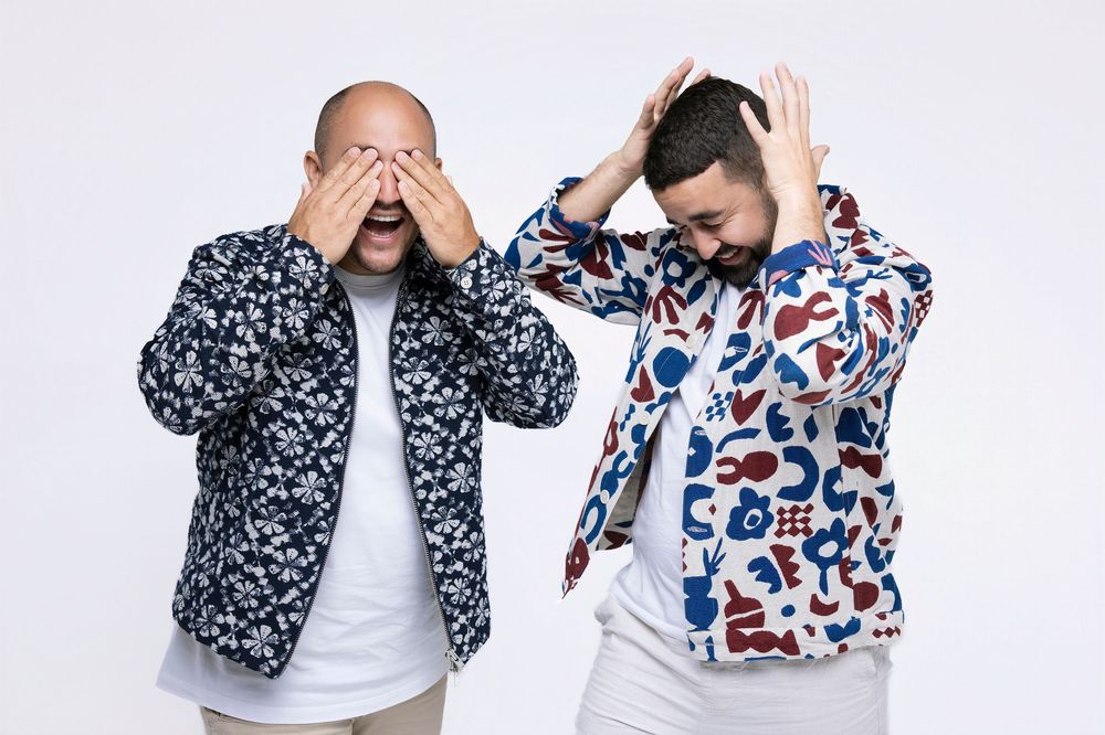 Two people in patterned bomber jackets covering their eyes while standing against a white background.