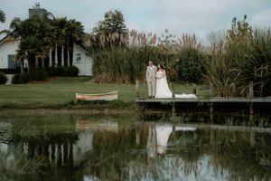 Bride and groom standing on a dock by a serene pond, with a white chapel and lush greenery reflected in the water.
