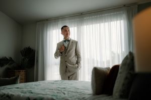 Groom in a light gray suit adjusting his green bow tie in a bright bedroom before the wedding ceremony.