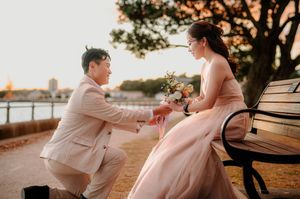 Couple in formal wear during romantic proposal on park bench at sunset, man kneeling with ring, woman in pink gown holding bouquet.