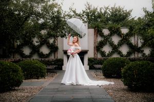 Bride in white gown holds clear umbrella in lush garden courtyard with manicured hedges and ivy walls.