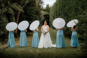 Bride in white gown poses with four bridesmaids in blue dresses holding white parasols in a lush garden setting.
