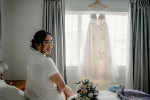 Bride smiling in white robe sitting on bed with wedding dress hanging in window and bouquet nearby.