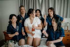 Bride in white robe with bridesmaids in navy pajamas celebrating with champagne glasses on a bed.