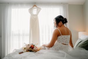 Bride sitting on bed in two-piece bridal outfit, gazing at white wedding dress hanging by window with floral bouquet nearby.