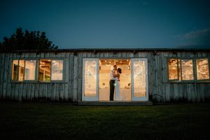 Couple embracing in lit barn doorway at dusk, romantic wedding venue with string lights glowing inside.