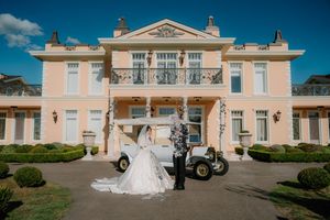 Bride and groom posing in front of a vintage white car and elegant pink mansion on a sunny wedding day.