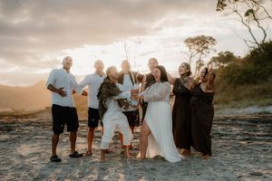 A joyful wedding party celebrates on a sandy beach at sunset, with two brides in white dresses surrounded by friends.