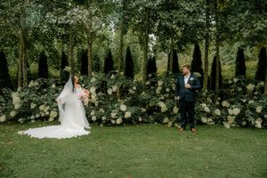 Bride and groom stand apart in lush garden with white hydrangea backdrop and tall trees at outdoor wedding ceremony.