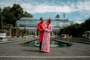 Couple in matching red traditional outfits posing by a garden fountain with a glass greenhouse backdrop.