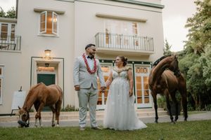 Bride and groom holding hands outdoors with two brown horses beside a elegant white venue building.