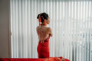 Woman in red qipao with intricate lace back detail stands by white blinds, elegant Chinese bridal fashion.