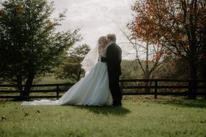 Bride and groom share a romantic kiss in a lush green field with autumn trees and rustic wooden fence backdrop.