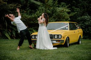 Bride and groom posing playfully in front of a vintage yellow station wagon on green grass at their wedding.