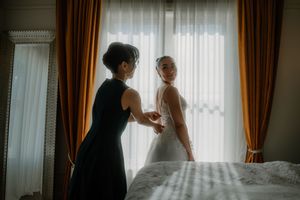A bridesmaid in black dress helps bride in white wedding gown get ready by a sunlit window with golden curtains.