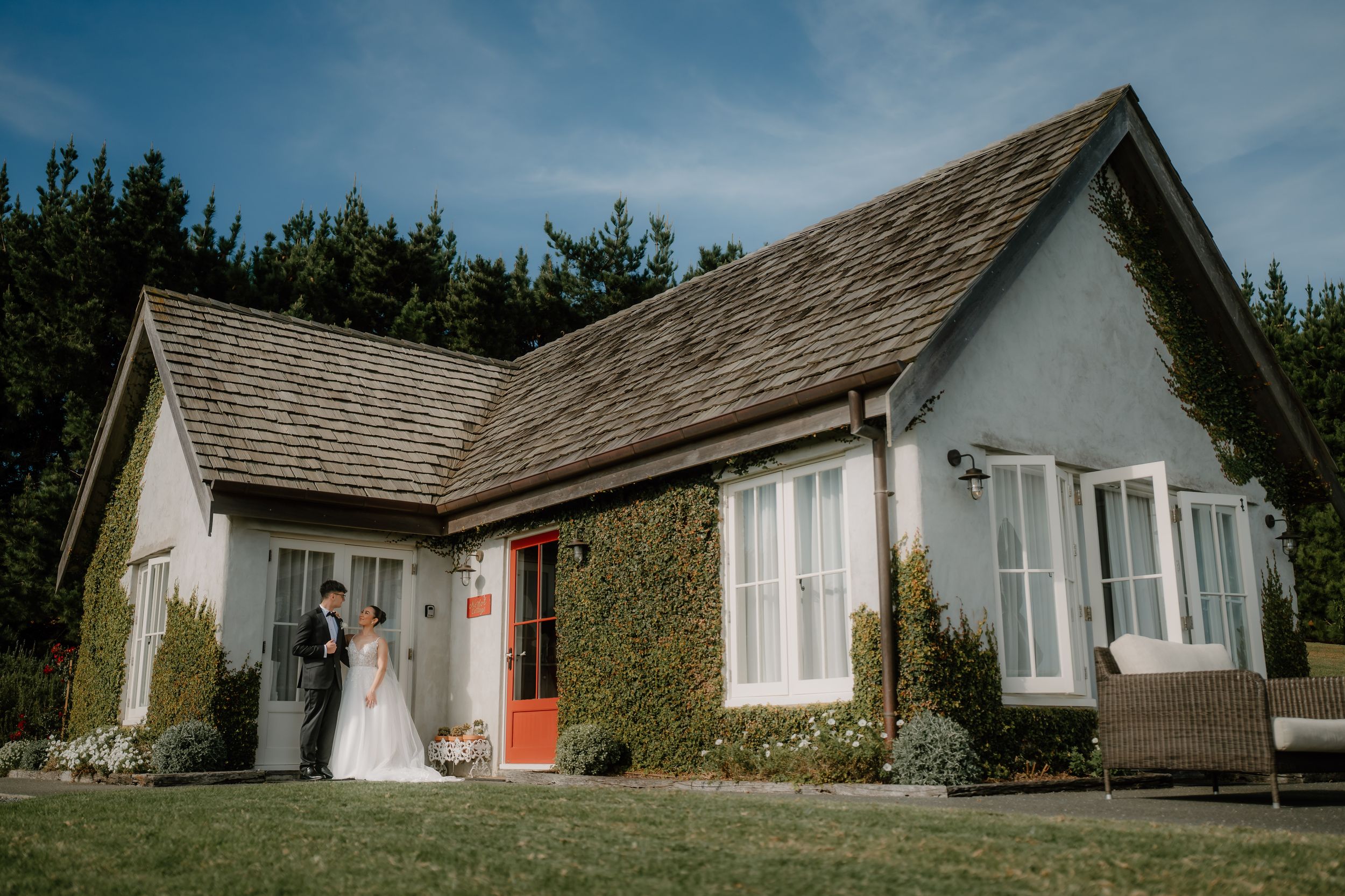 Bride and groom posing outside a charming white cottage wedding venue with ivy-covered walls and a red door.