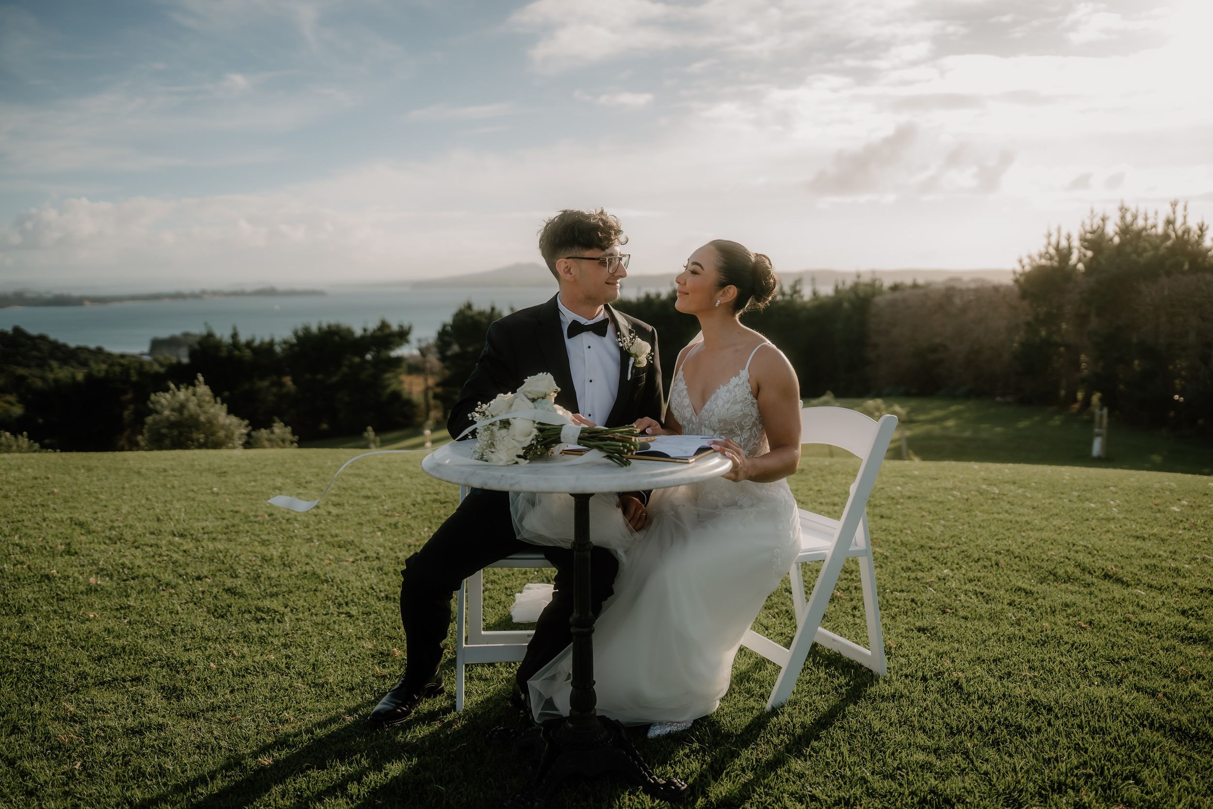 Bride and groom sitting at a small white table on a green hillside with ocean views on their wedding day.