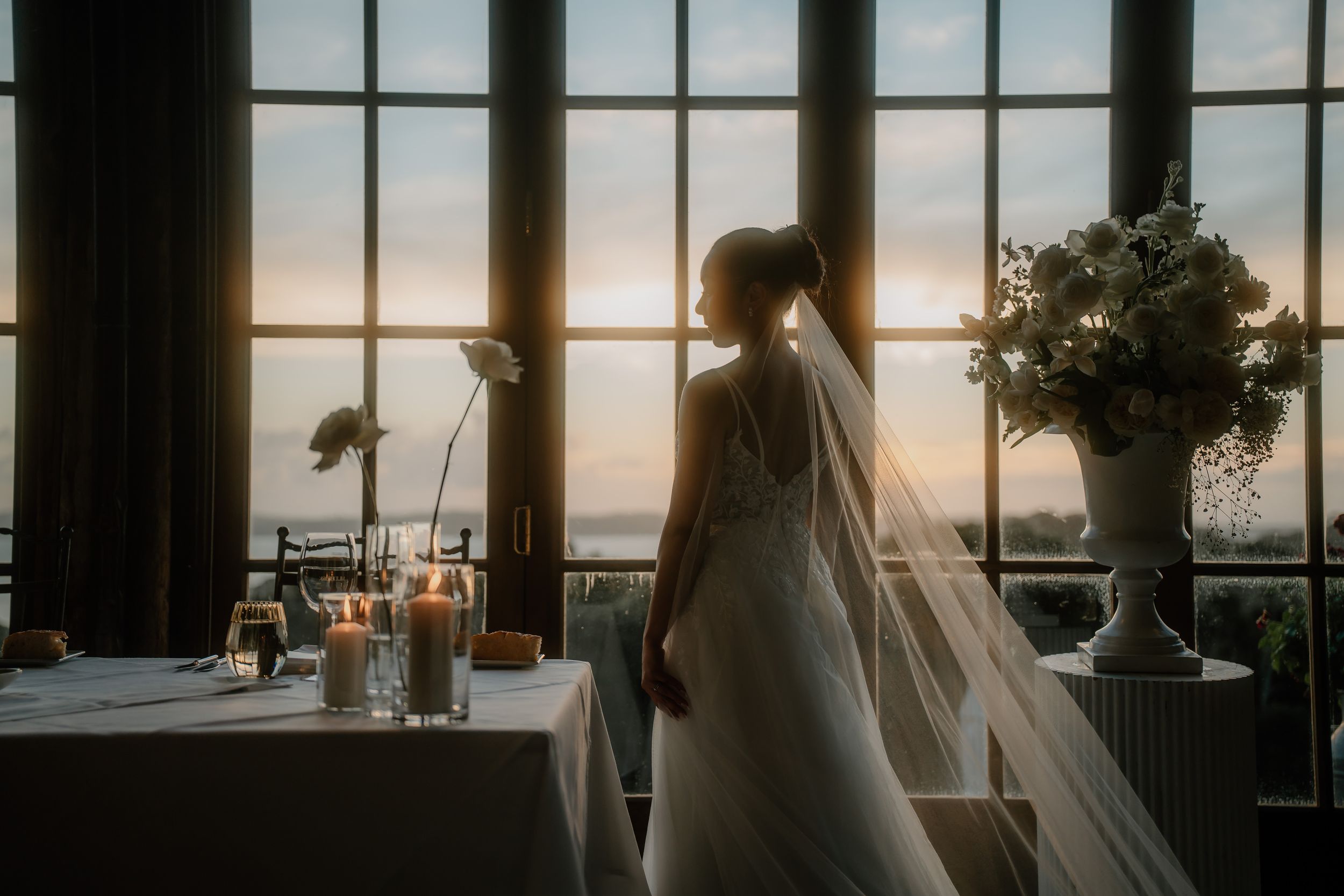 Bride in elegant gown and veil stands by large windows at golden sunset, with candles and floral arrangements nearby.