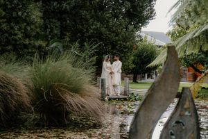 Couple in white robes standing on a bridge over a lily pond surrounded by lush greenery in a serene garden setting.