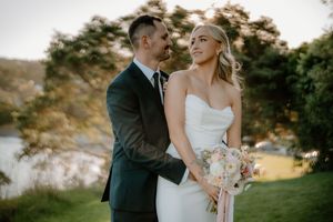 Bride holding floral bouquet stands with groom in dark suit outdoors near waterfront at golden hour wedding.