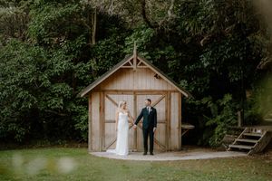 Bride and groom holding hands in front of a rustic wooden chapel surrounded by lush green trees.