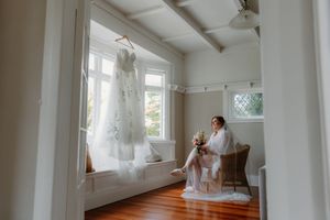 Bride in white robe sits in chair holding bouquet beside hanging wedding dress in bright room.