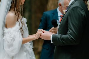 Bride and groom exchange rings during outdoor wedding ceremony, officiant holding microphone in background.