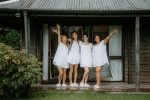 Four women in white dresses pose joyfully on a rustic wooden porch, arms raised in celebration.