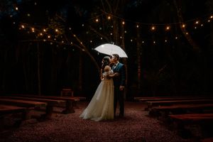 Bride and groom share romantic kiss under white umbrella at outdoor woodland wedding venue with string lights at night.