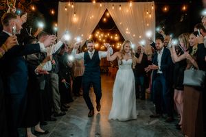 Joyful newlyweds exit wedding reception through sparkler sendoff tunnel with guests celebrating under warm string lights.