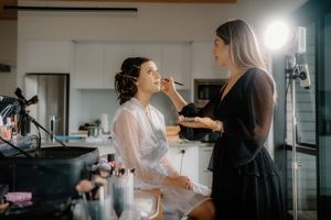Makeup artist applying makeup to a bride in a white robe before her wedding day in a bright studio.