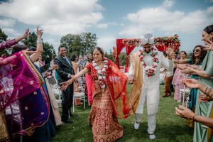 Indian bride and groom walk down the aisle at outdoor wedding ceremony, surrounded by colorful guests celebrating.
