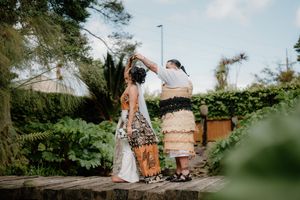 Two women in traditional Polynesian attire share a ceremonial moment outdoors, surrounded by lush greenery.