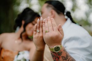 Newlyweds displaying wedding rings on tattooed hands with a Rolex watch, couple kissing softly in background.