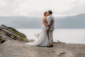 Bride and groom share a romantic kiss on rocky cliffside overlooking misty lake and mountains on wedding day.