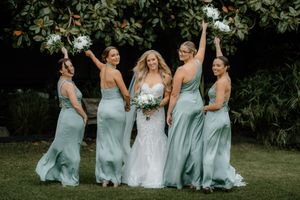 Bride in white lace gown with four bridesmaids in sage green dresses holding bouquets outdoors.