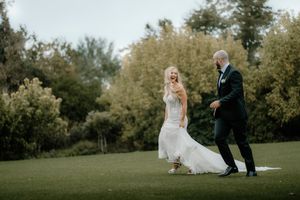 Bride and groom walking on lush green lawn surrounded by trees during outdoor wedding celebration.