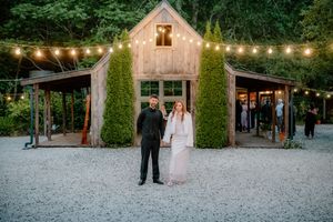 Couple holding hands in front of a rustic barn venue with string lights and lush greenery at twilight wedding reception.