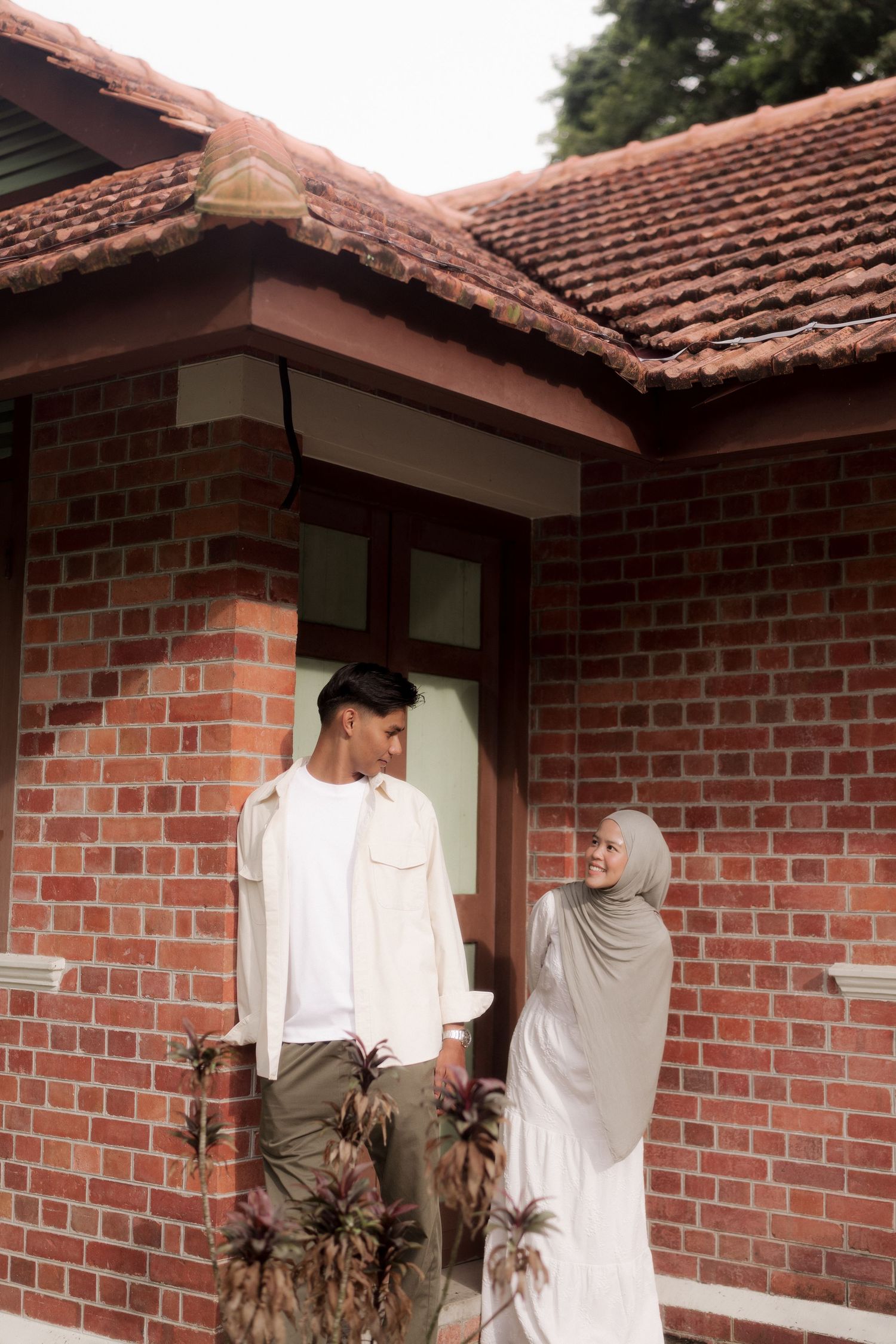 A Muslim couple stands together outside a red brick building with terracotta roof tiles, smiling at each other.
