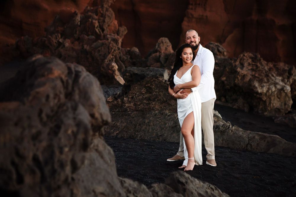 Couple embracing on black volcanic rocks at a scenic coastal location, woman in white dress, man in white shirt.