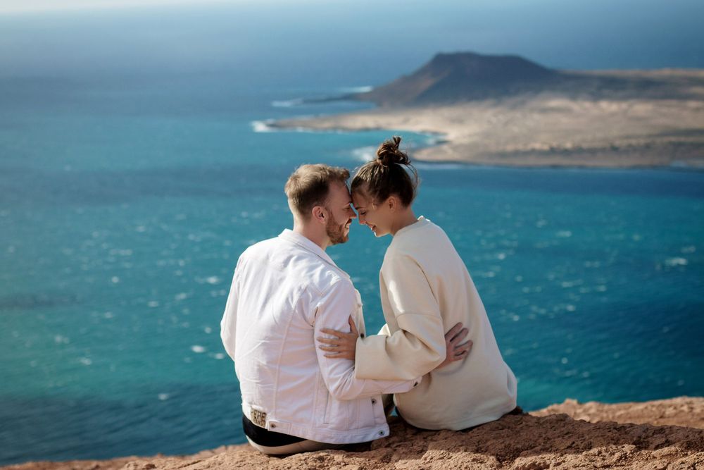 Romantic couple sitting on a cliff edge sharing an intimate moment with turquoise ocean and island views behind them.