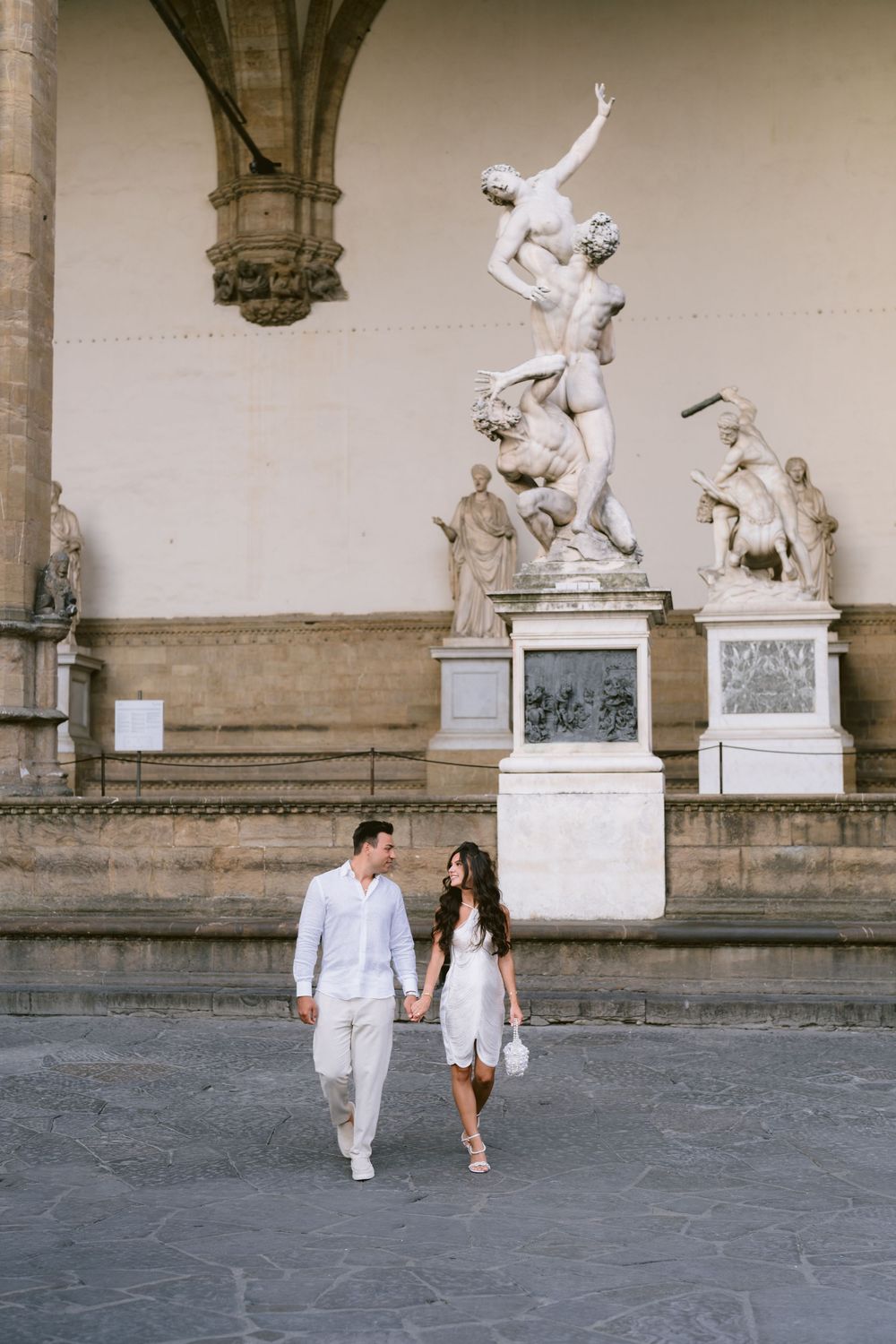 Couple holding hands walking in front of classical sculptures at Piazza della Signoria, Florence, Italy.