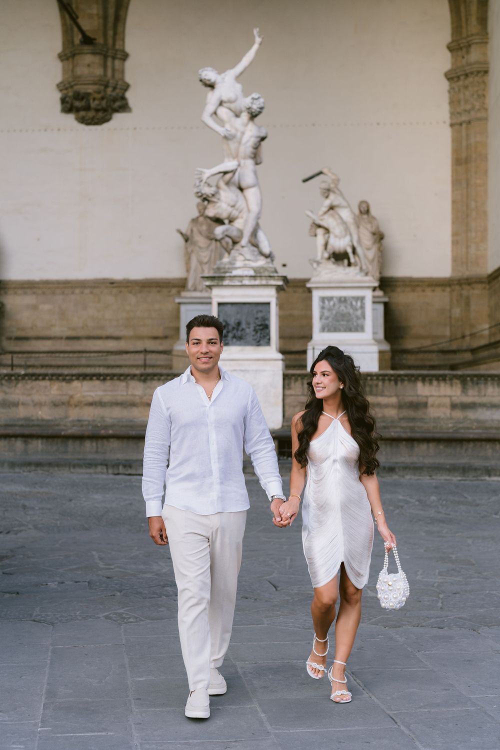 Couple in white outfits holding hands at Piazza della Signoria, Florence, with classical sculptures behind them.