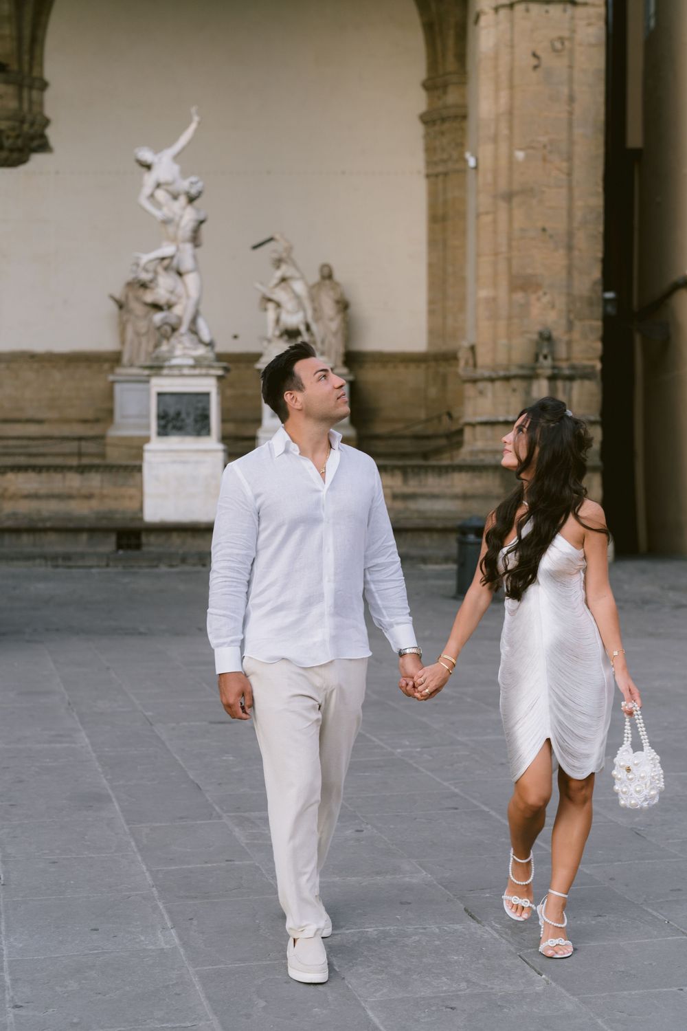 Couple in white outfits holding hands at Piazza della Signoria, Florence, with classical sculpture backdrop.