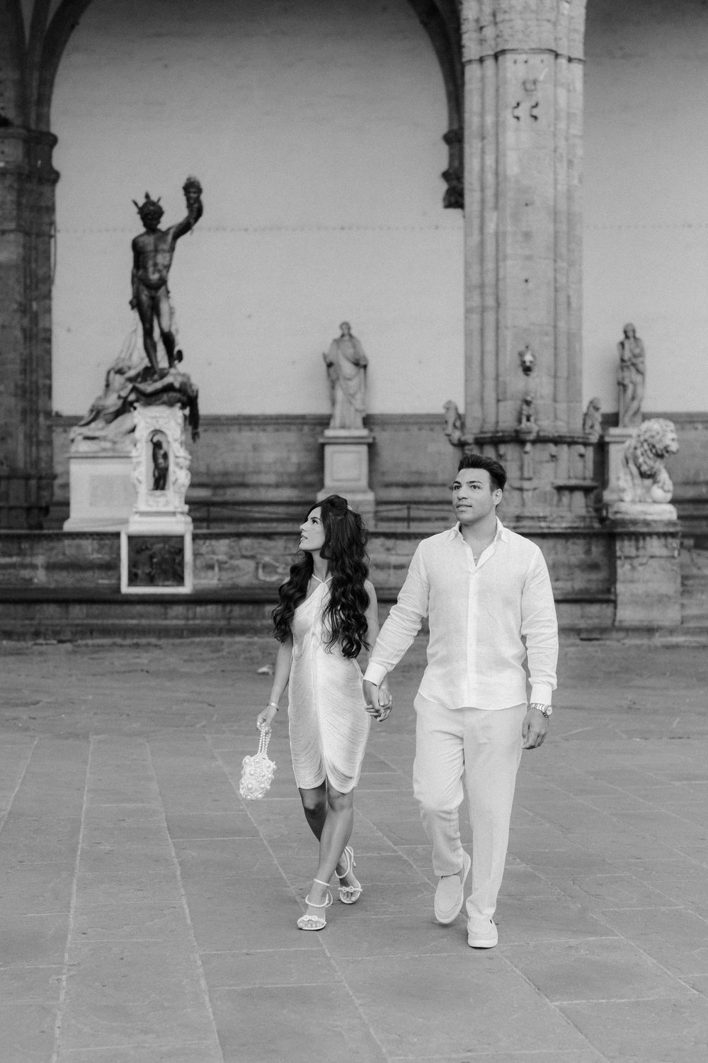 Black and white photo of a couple holding hands walking past classical sculptures at Piazza della Signoria, Florence.