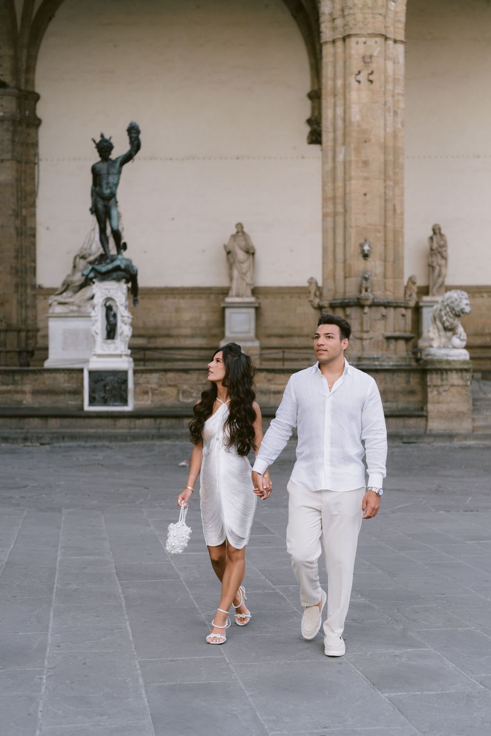 Couple in white outfits holding hands walking in front of classical statues at Loggia dei Lanzi, Florence.