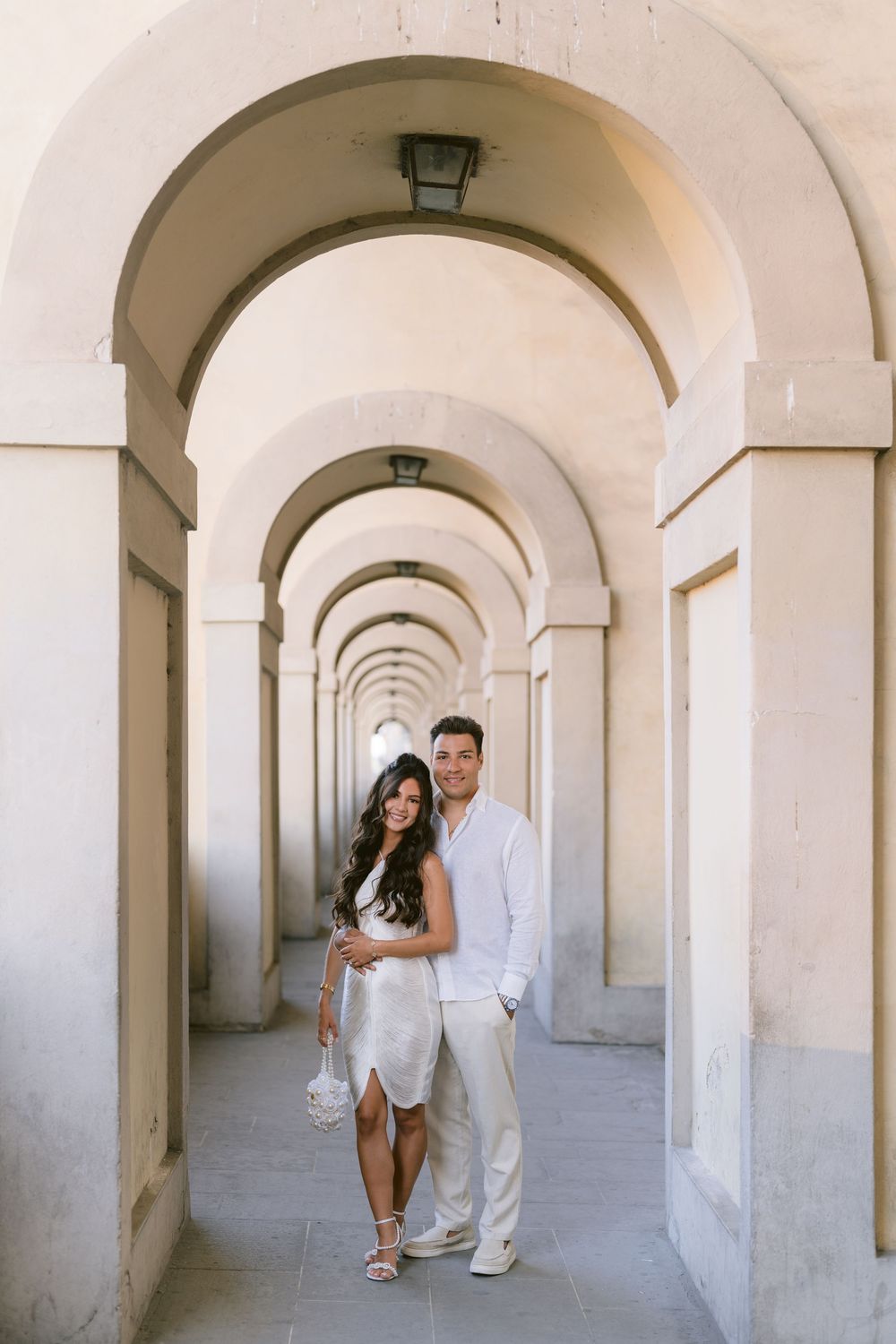 Couple in white outfits posing under elegant arched colonnade corridor, romantic engagement photo session.