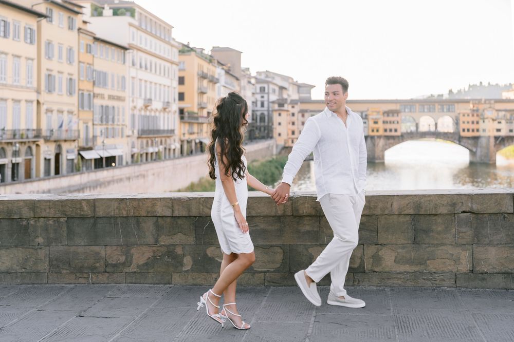 Couple holding hands walking along the Arno River with Ponte Vecchio in Florence, Italy in background.